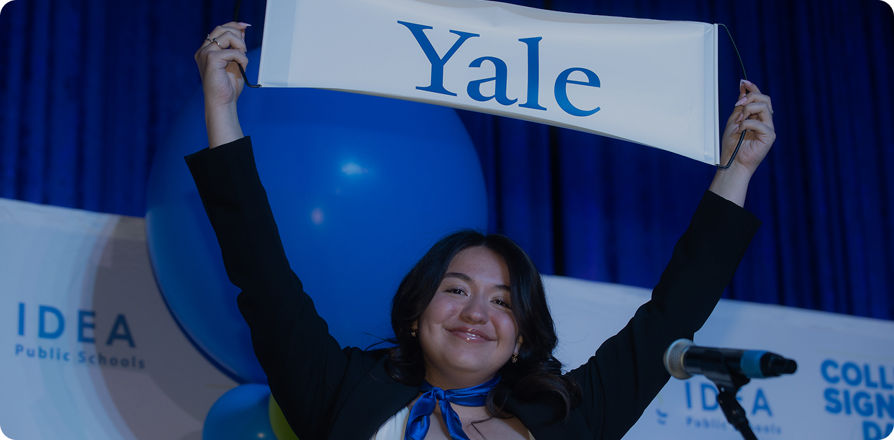 A smiling student proudly holds up a Yale banner overhead during a college signing celebration, with blue and gold balloons and an IDEA Public Schools backdrop behind her